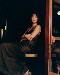 Portrait of actress Lizzy Caplan wearing a dark sheer dress seated indoors near a reflective wooden frame mirror.
