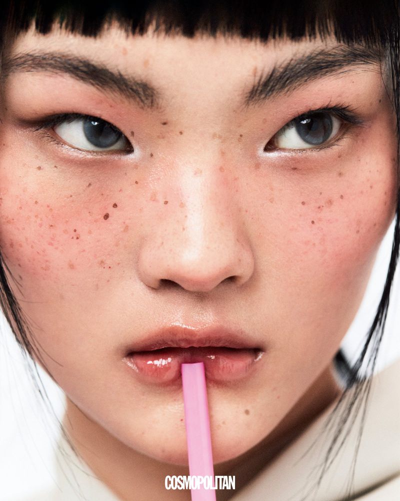 Close-up of a young Asian woman with a pink straw in her mouth, light eyes, freckles, and dark bangs, with the 'Cosmopolitan' logo.