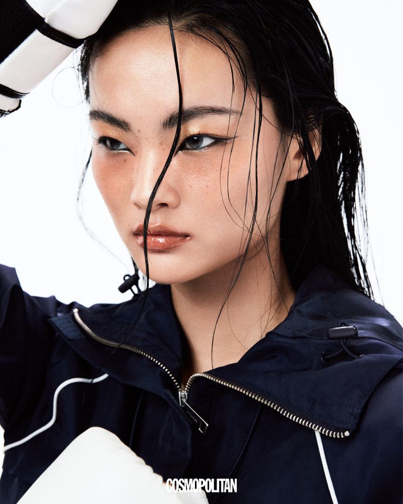 East Asian woman with wet hair wearing a navy athletic jacket and white boxing gloves against a white studio background.
