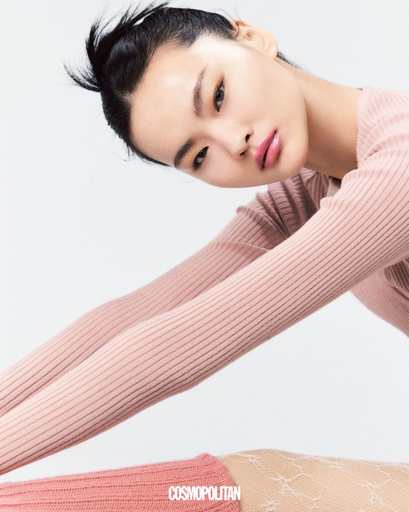 Asian woman in a pink ribbed sweater posing in a photo studio. She has her hair in a bun, light pink lip color, and is smiling.