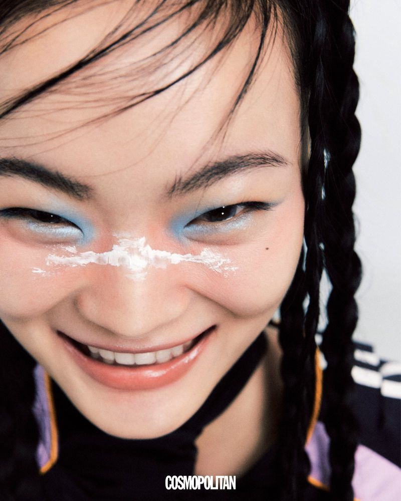 Close-up of a smiling Asian woman with creative makeup and braided hair, photographed in a light studio environment for Cosmopolitan.