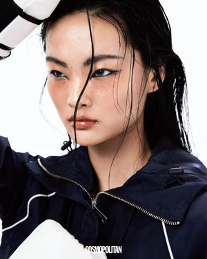 East Asian woman with wet hair wearing a navy athletic jacket and white boxing gloves against a white studio background.