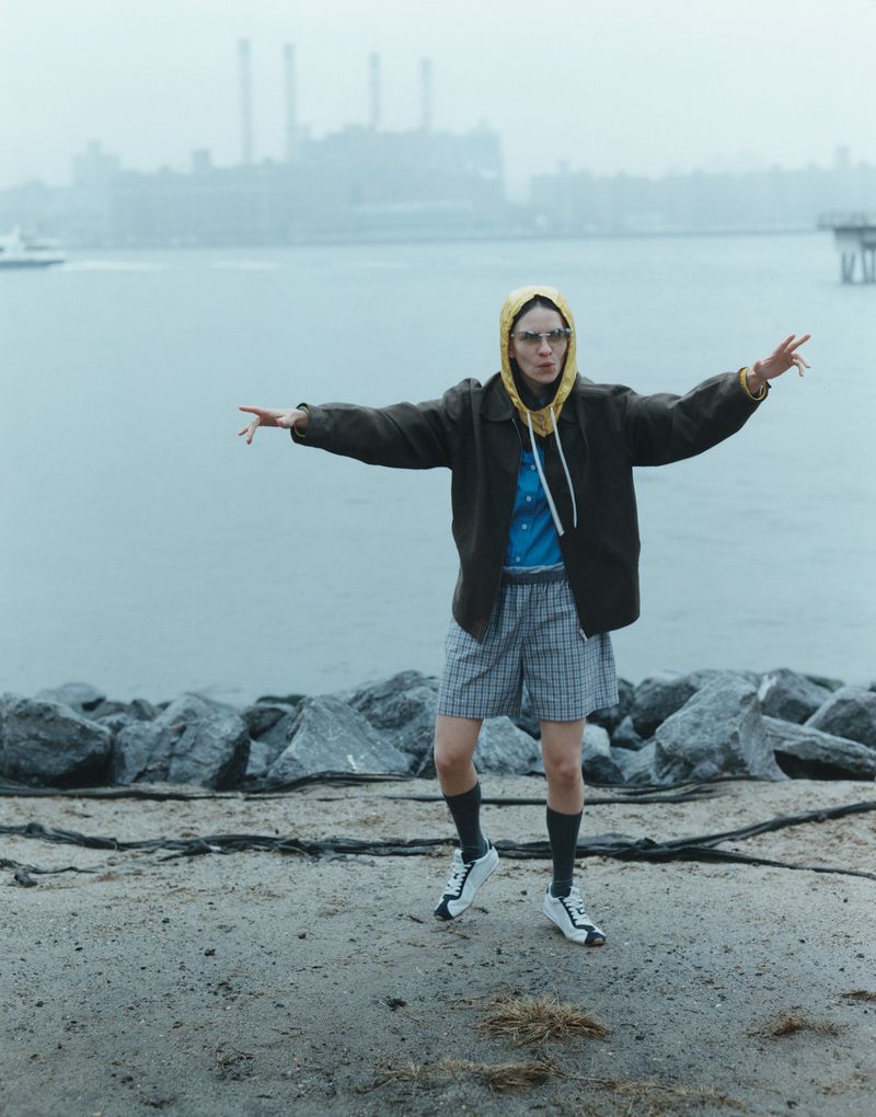Adult female with arms outstretched on a rocky beach. Wearing shorts, jacket, glasses, and sneakers. Water and buildings in background.