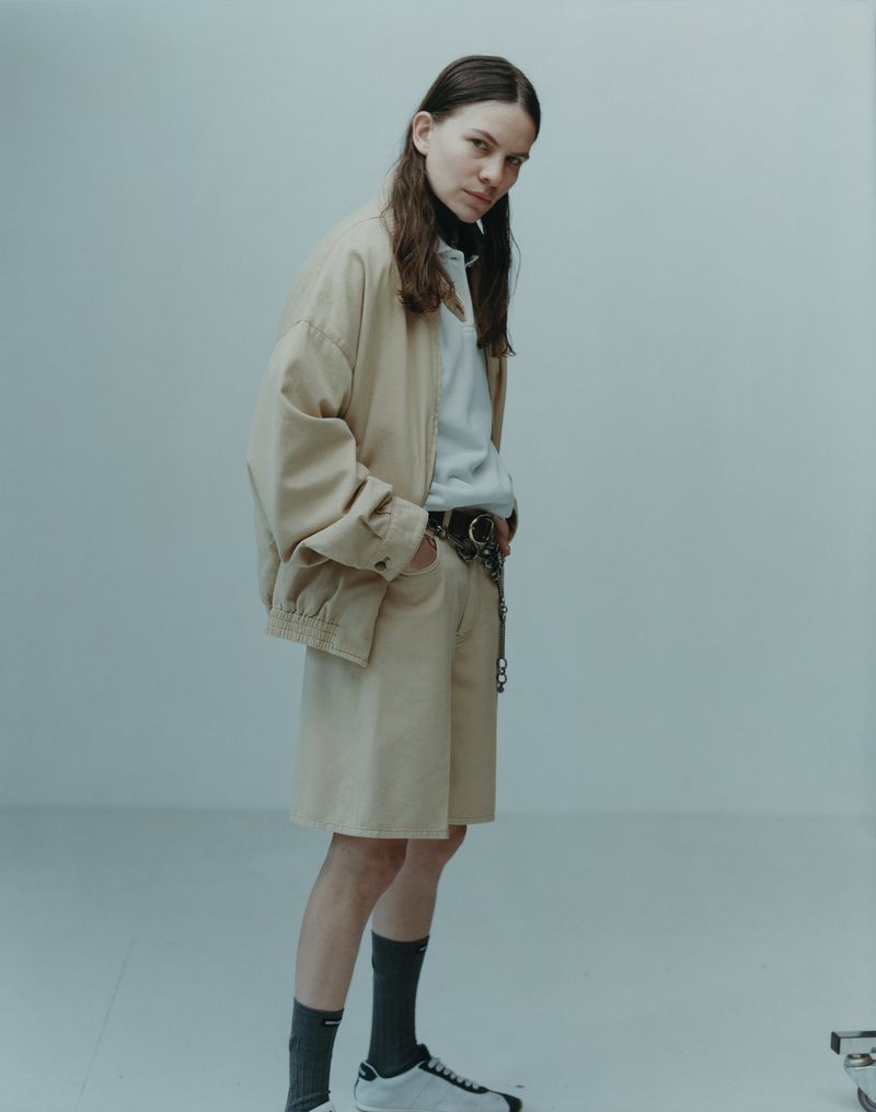 Full-length portrait of a model in a beige jacket and shorts, wearing sneakers against a neutral backdrop in a studio setting.