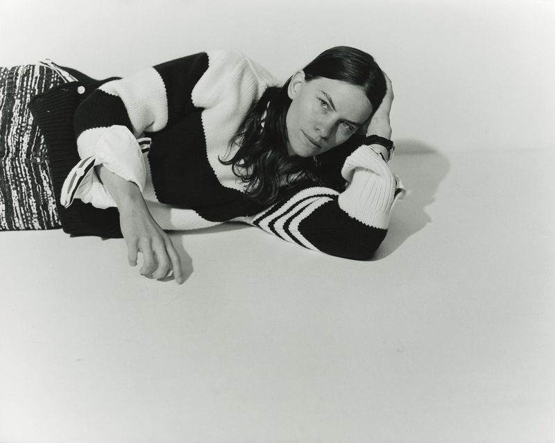 Monochrome portrait of a woman reclining on a white surface, wearing a patterned sweater and shirt. Studio lighting emphasizes the subject.