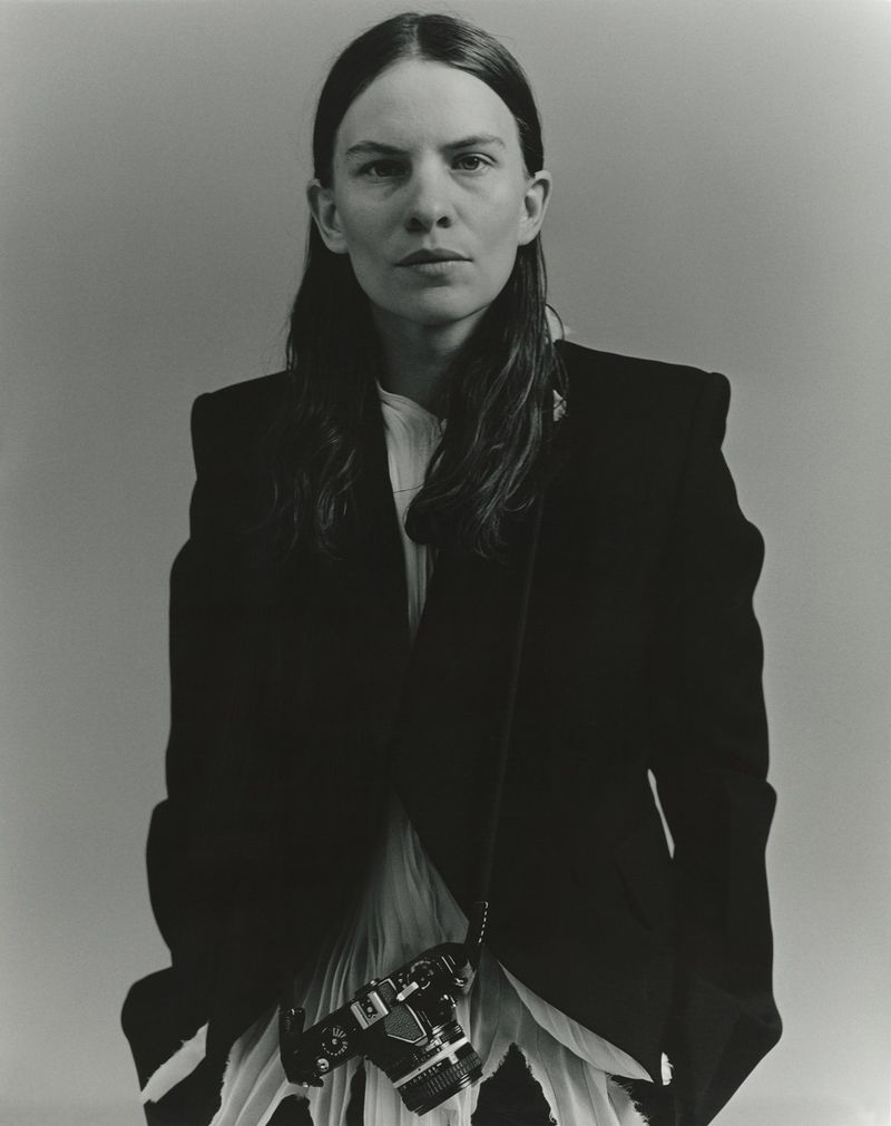 Monochrome portrait of a woman with long dark hair wearing a black blazer and pleated skirt, holding a camera against a grey backdrop.