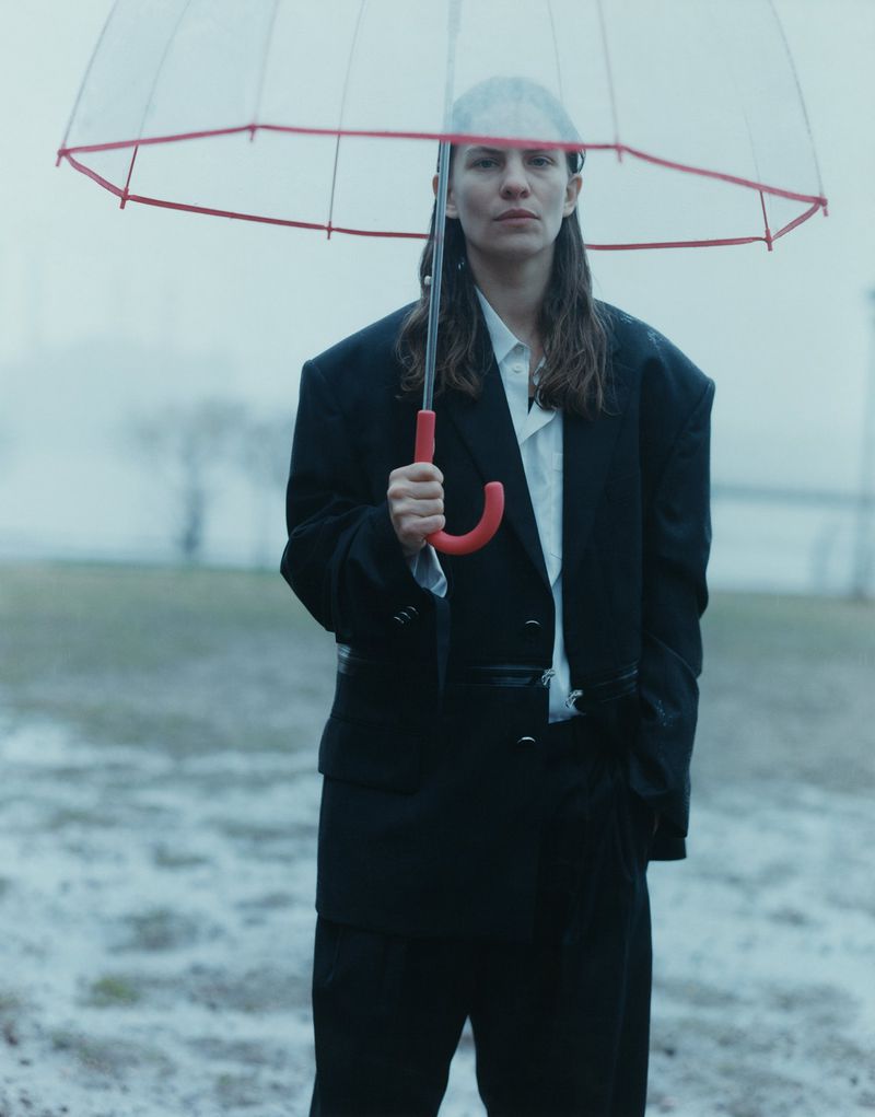 Woman in a black suit stands outside holding a clear umbrella on a rainy day. She has long hair and looks directly at the camera.