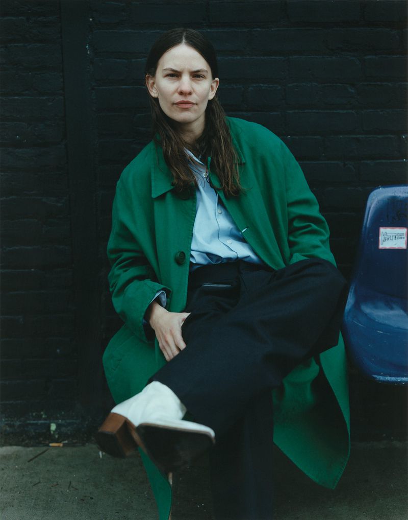 Portrait of a woman with long hair wearing a green coat, sitting outdoors against a dark brick wall, looking directly at the camera.