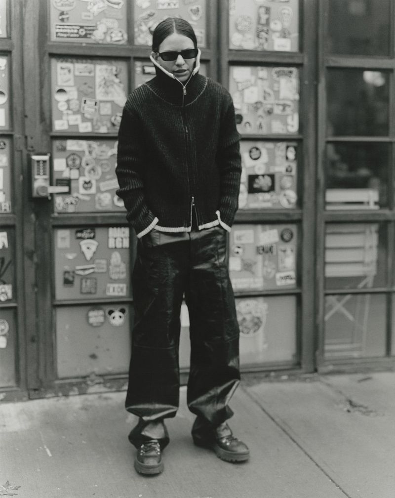 A person in a black jacket and baggy pants stands in front of a sticker-covered glass door in a monochrome portrait-style shot.