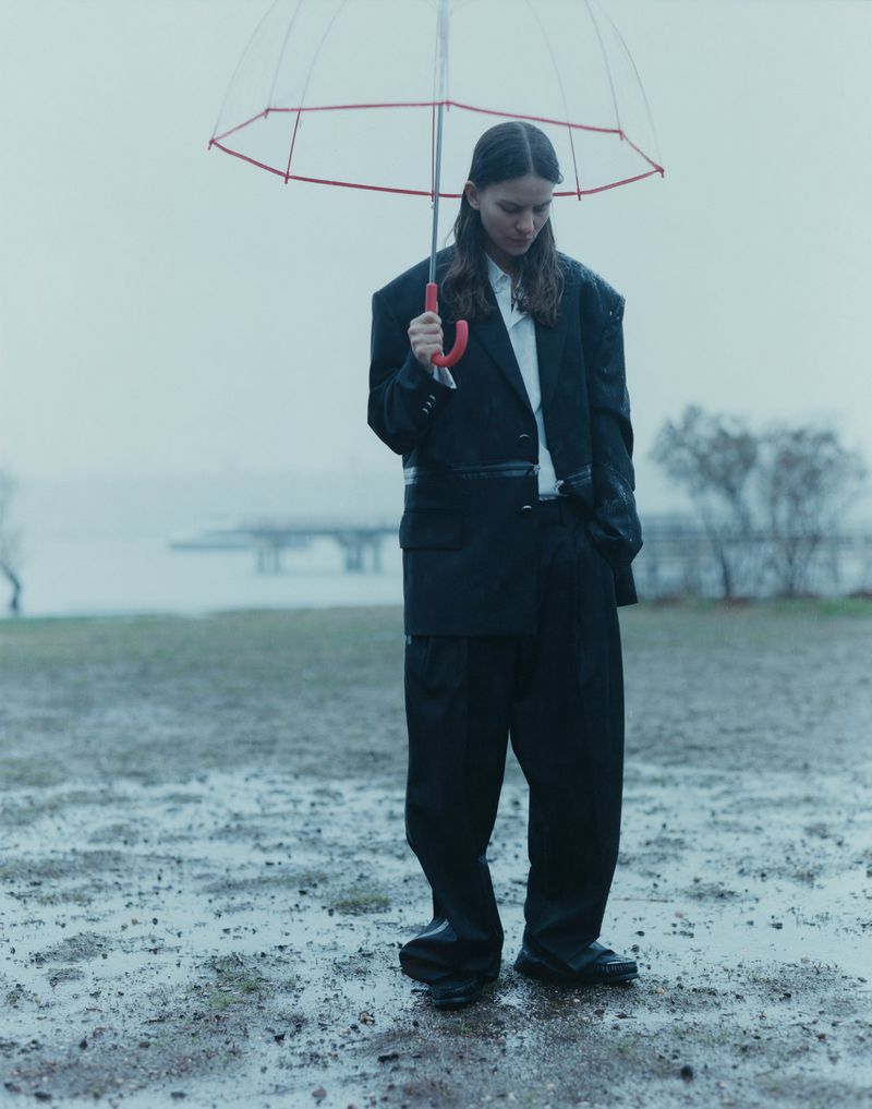 Woman in oversized blazer and trousers stands under clear umbrella in rainy outdoor setting near a body of water.