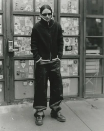 Eliot Sumner wearing a zip-up knit jacket, dark wide-leg trousers, and sunglasses in front of sticker-covered glass doors.