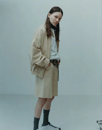 Eliot Sumner posing in an oversized beige jacket and matching shorts in a minimalist studio for METAL Magazine.