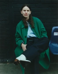 Eliot Sumner seated wearing an emerald green overcoat, black trousers, and white heeled boots against a dark brick wall.