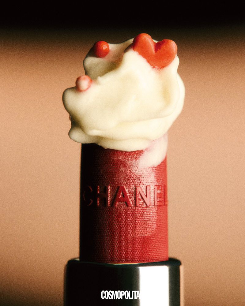 Close-up of bright red Chanel lipstick with white cream and red heart candies on top, set against a soft peach background.
