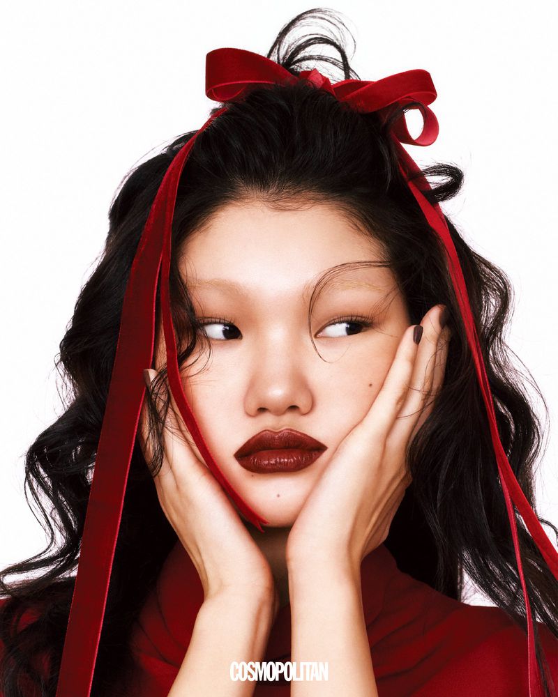 An Asian female model with red lips, dark hair, and a velvet red bow poses against a white background, in a beauty fashion-style portrait.