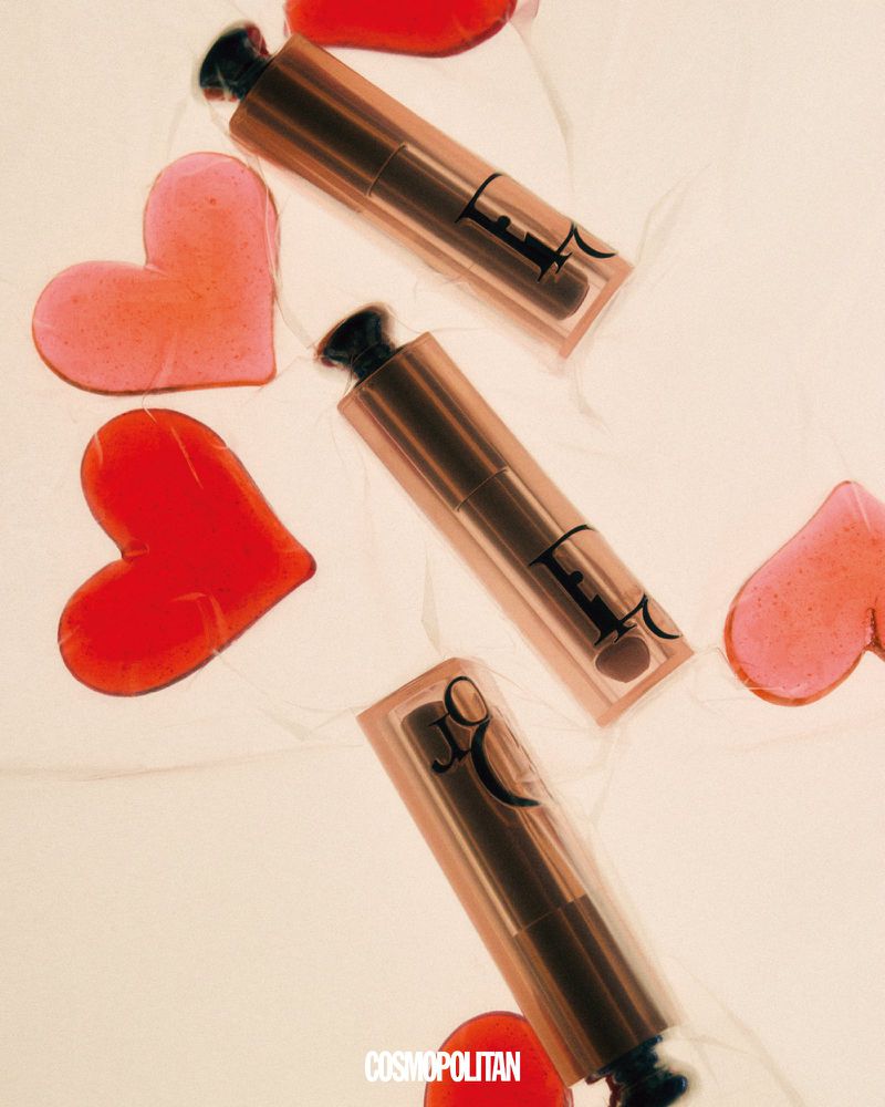 Cosmetics flat lay: three rose-gold lipsticks with heart-shaped accents on a light, textured background. Beauty product photography.