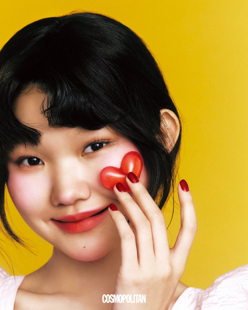Close-up portrait of a young Asian woman with short dark hair, a red heart on her cheek, bright red lips and nails, on yellow backdrop.