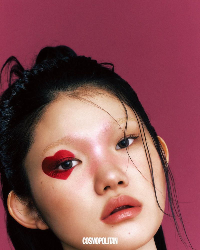 Close-up portrait of an Asian woman with heart-shaped makeup on her eye, set against a solid dark-pink backdrop in studio lighting.