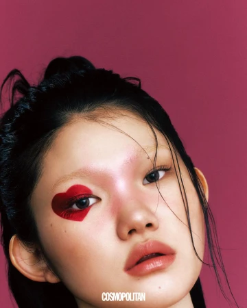 Close-up beauty portrait of an East Asian woman with a bold red heart painted around her eye against a magenta backdrop.