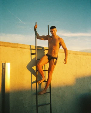 Model Christopher Lima in black swimwear climbing a vertical metal ladder on a sunlit rooftop wall.