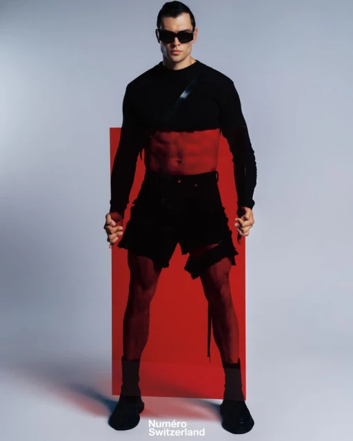 Fashion photo of a muscular male model in black two-piece outfit and sunglasses, posing against a neutral background with red prop.