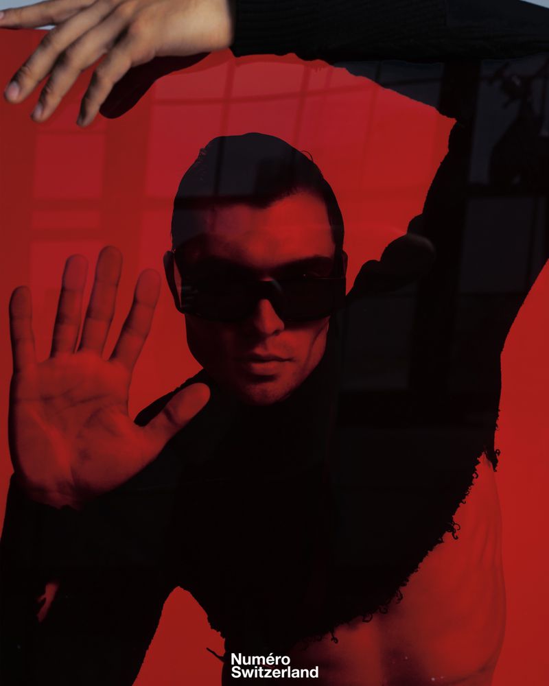 Portrait of a man with sunglasses against a red background, hands visible, wearing a black turtleneck sweater, studio shot.