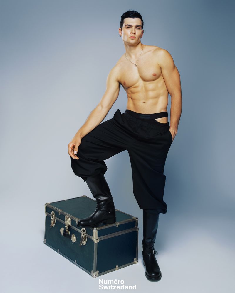 Male fashion model poses shirtless in black pants and boots, with a dark green trunk against a light blue studio background.