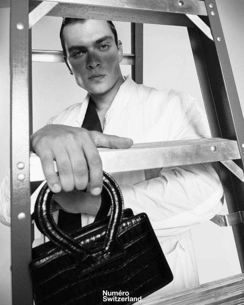 Model Kirill Kabachenko poses behind a metal ladder holding a black crocodile-embossed handbag against a studio backdrop.