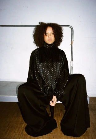 Model with voluminous curly hair seated wearing an oversized black polka-dot top and wide-leg trousers against a white wall.