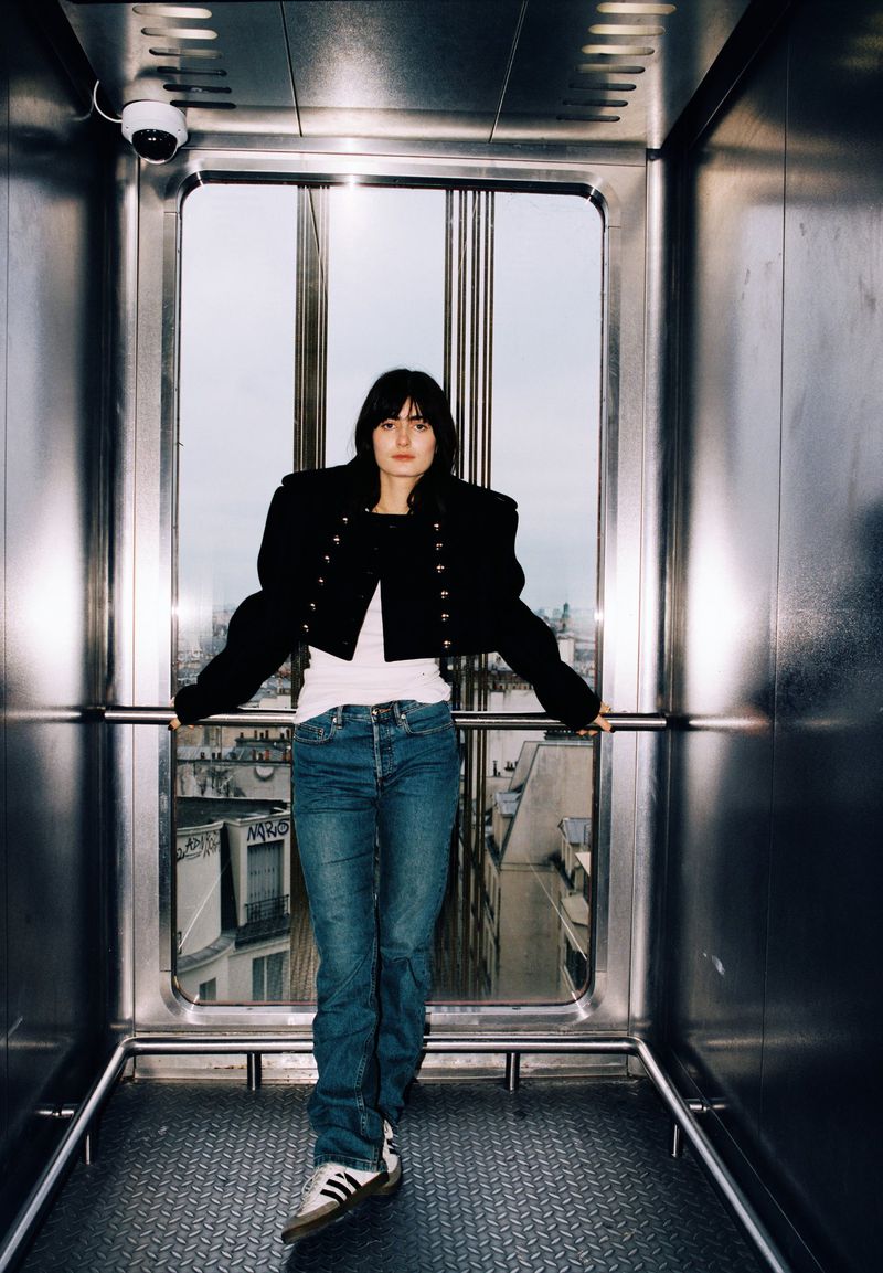 Young woman poses in an elevator with a city view. She wears a black jacket, white shirt, jeans, and Adidas sneakers in an indoor setting.