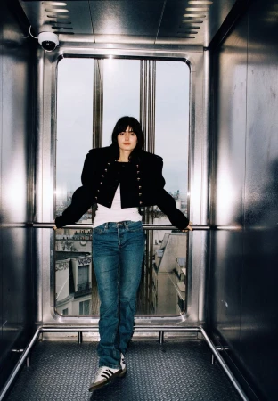 Young woman poses in an elevator with a city view. She wears a black jacket, white shirt, jeans, and Adidas sneakers in an indoor setting.