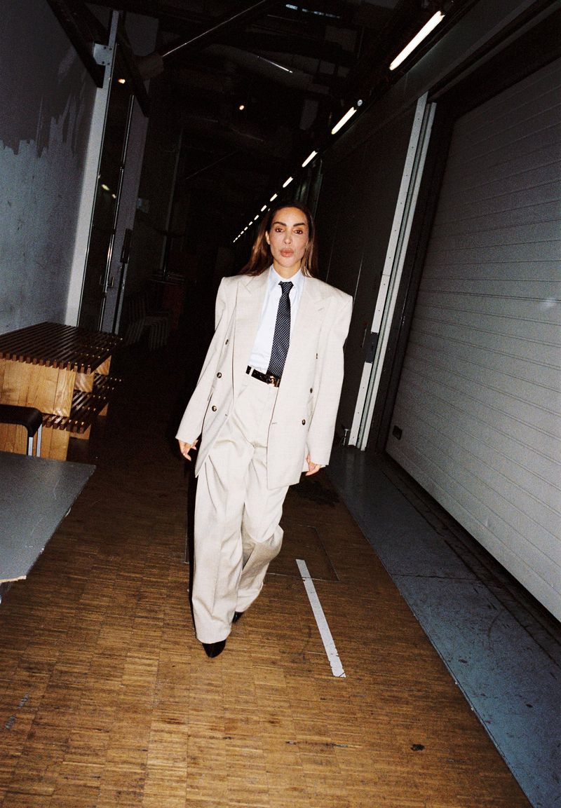A woman in an oversized beige suit and tie stands on a wooden floor in a dimly lit hallway with bright lights overhead.