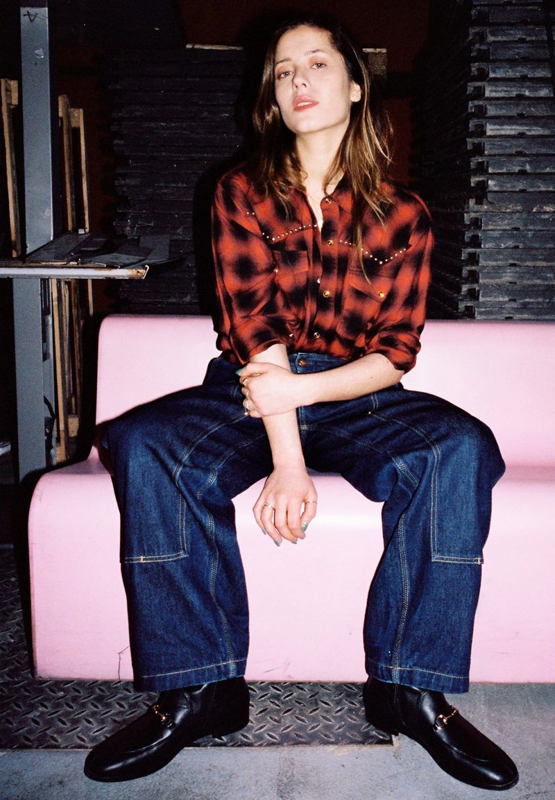 A brunette woman poses in a red plaid shirt and baggy blue jeans, sitting on a pink couch in a casual, indoor studio setting.