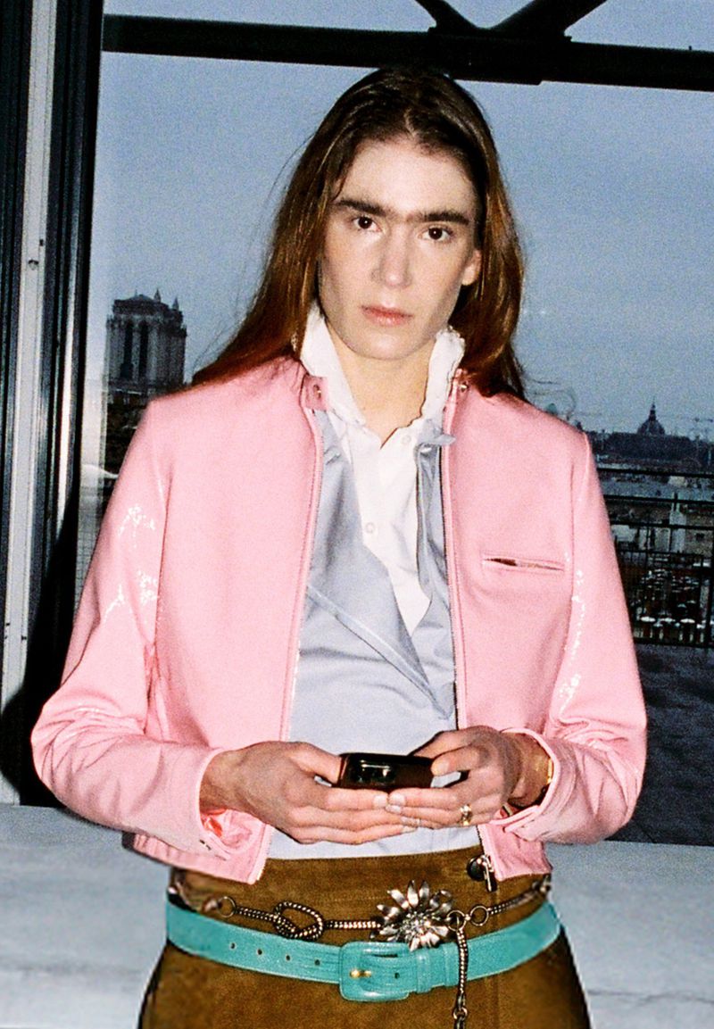 Young adult with long hair wears a pink jacket and turquoise belt, holding a device, with Paris buildings visible through the window.