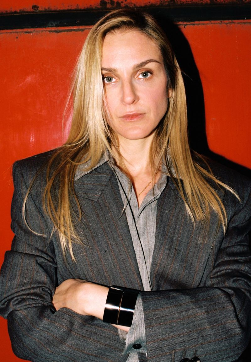Portrait of an adult woman with long blonde hair wearing a grey blazer and a black cuff bracelet against a solid red background.