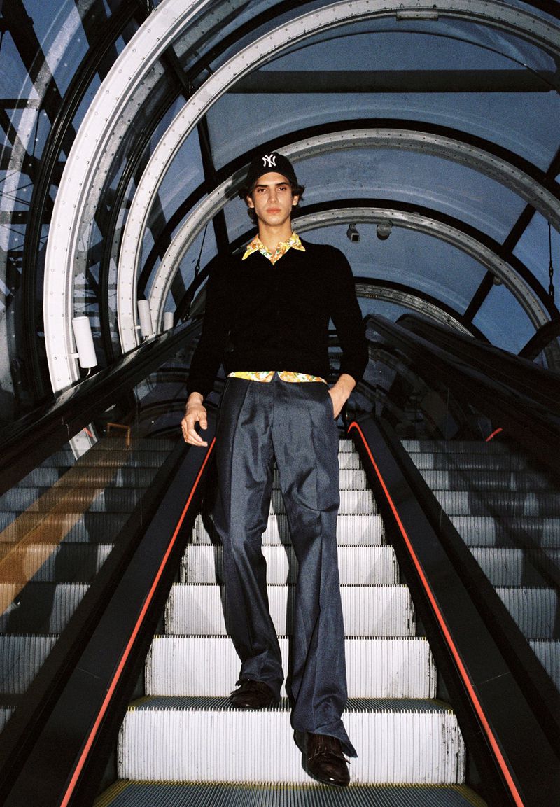 A young adult man wearing a baseball cap, yellow shirt, and blue slacks, posing on an escalator under a glass arched ceiling.