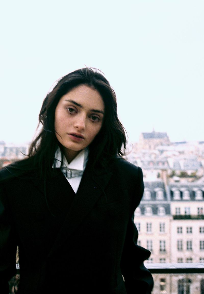 Model wearing a black tailored blazer and white collared shirt on a balcony overlooking Parisian rooftops.