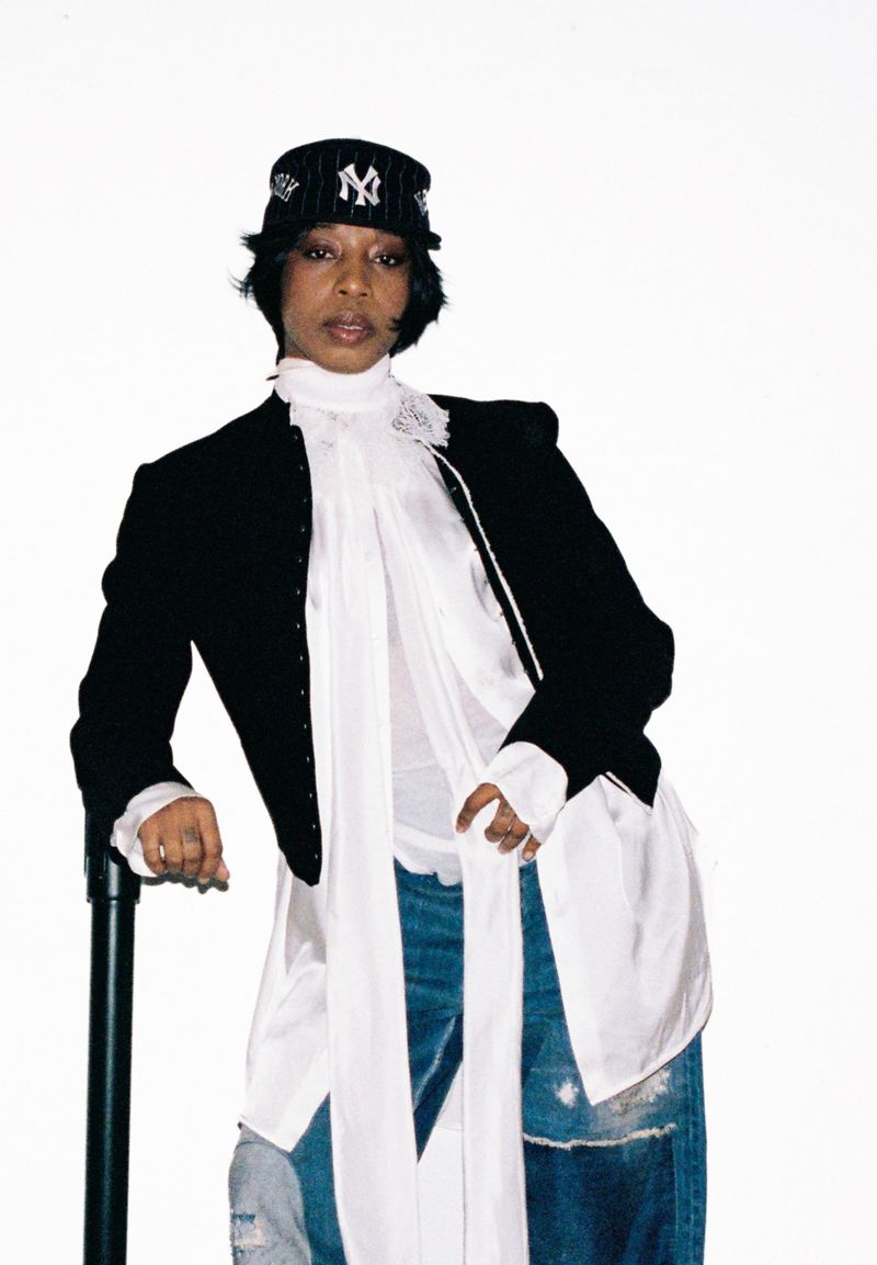 Adult female poses indoors wearing a baseball cap, blazer, long white shirt, and patchwork jeans leaning on a black pole.