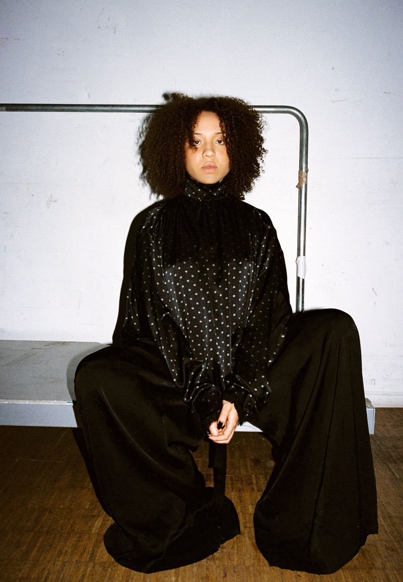 Model with voluminous curly hair seated wearing an oversized black polka-dot top and wide-leg trousers against a white wall.