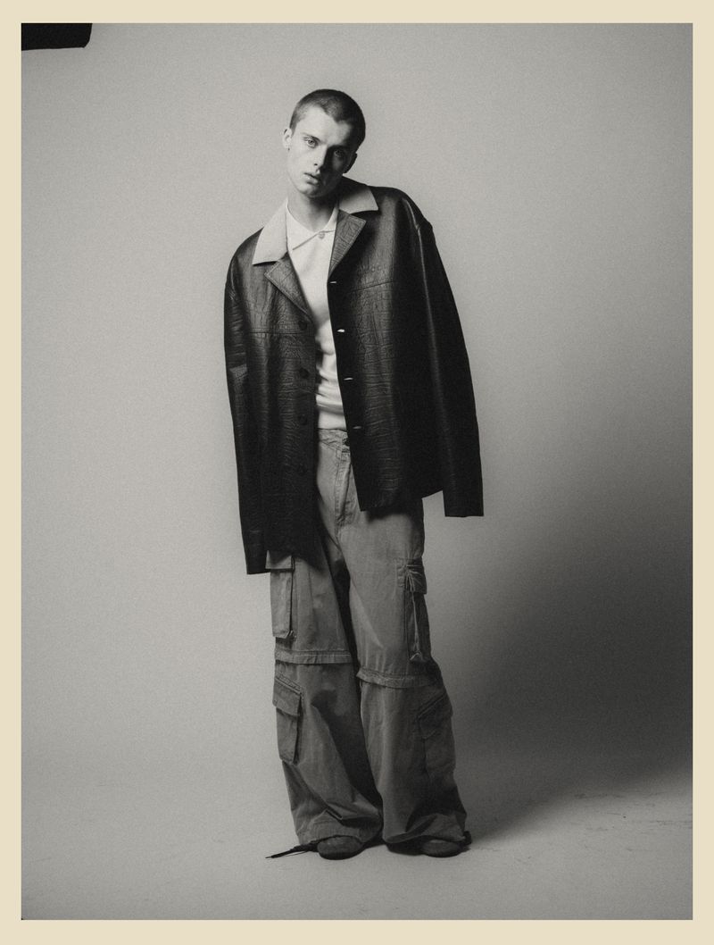 Full-length studio shot of a young adult male in a leather jacket, white polo shirt, and khaki cargo pants against a gray backdrop.