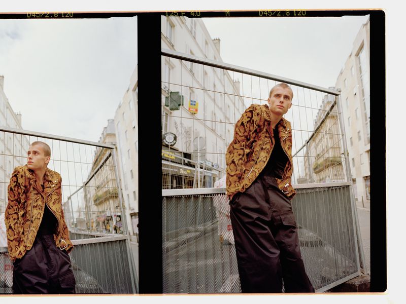 Adult male poses in patterned jacket and dark pants leaning against a metal barricade on an urban street with buildings.