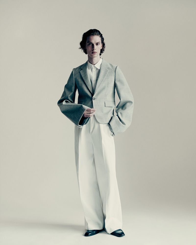 A stylish young adult male stands in a photo studio. He's wearing a light green blazer with wide sleeves and white trousers and black shoes.