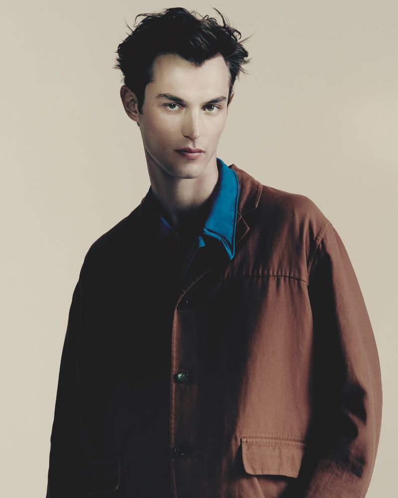 Portrait of a young adult male fashion model in a brown jacket and a blue collared shirt, posing indoors against a neutral background.