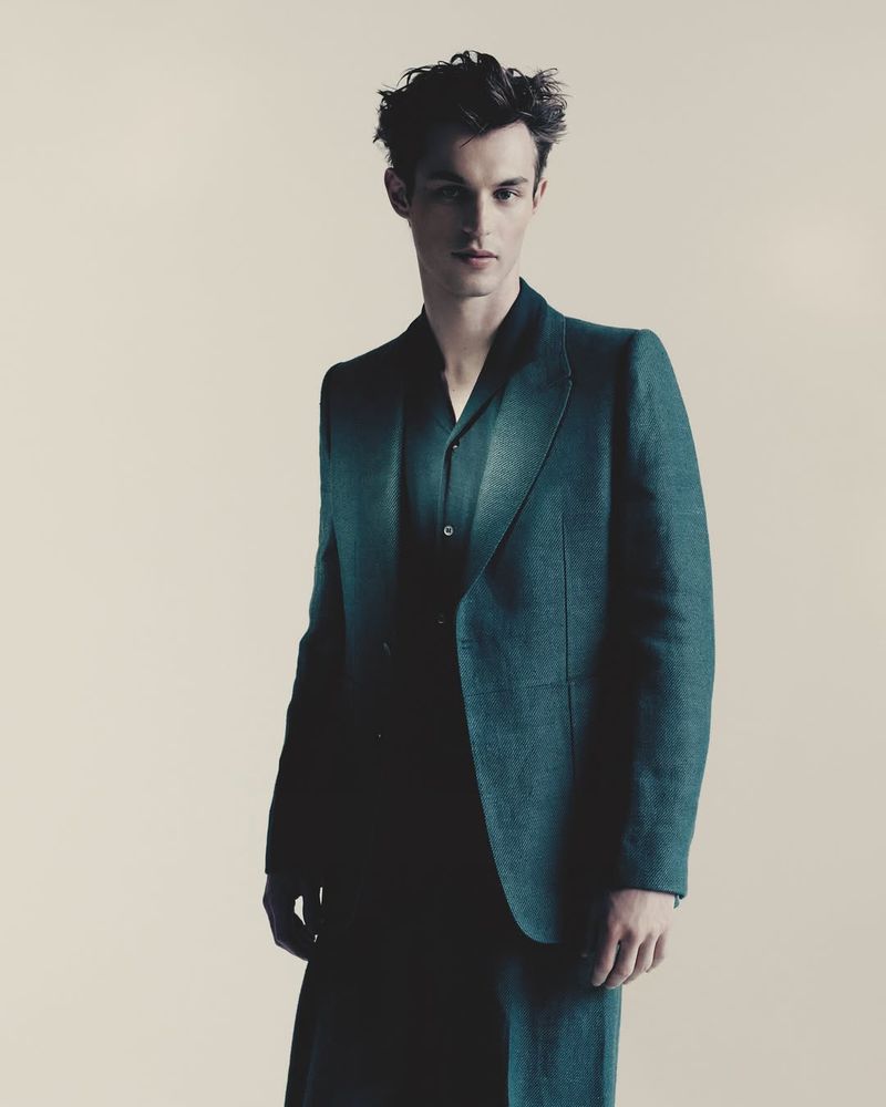 Kit Butler posing in a tailored dark green suit against a warm neutral studio backdrop.
