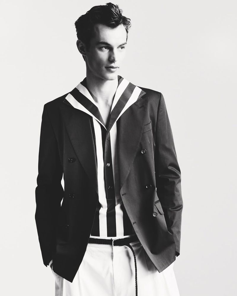 Monochrome fashion shot of a young adult man in a dark blazer, striped shirt, and light-colored trousers in a minimalist studio setting.