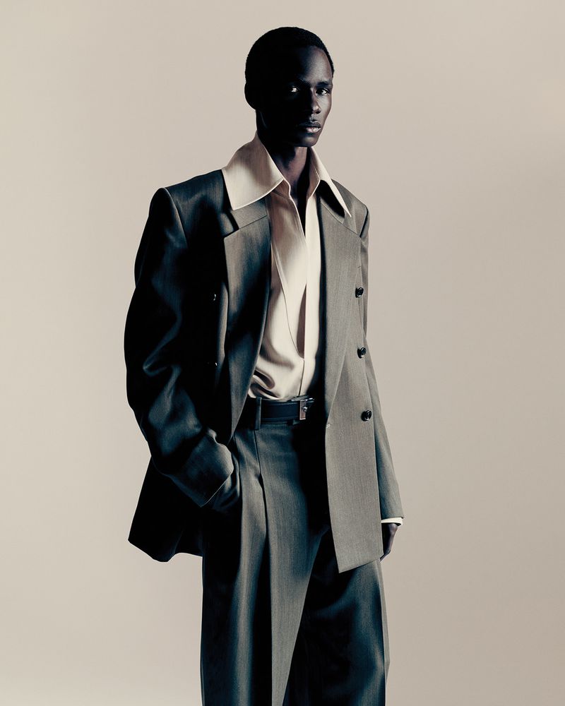 Editorial fashion shot of a black male model wearing a gray oversized suit with wide lapels, and flared pants, posing in a studio.