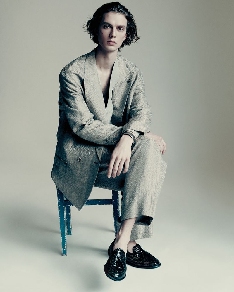 Young adult male in a grey textured suit, seated on a blue chair in a photo studio setting, wearing black loafers and a bracelet.