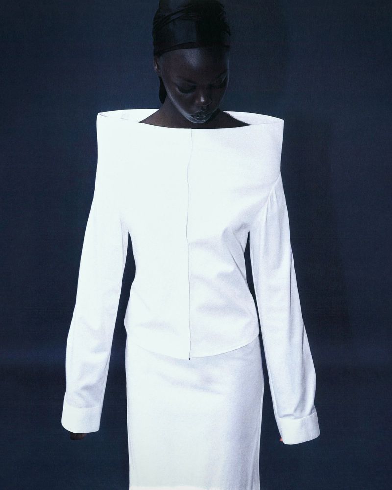Fashionable woman in white tailored skirt suit with unique shoulders and head wrap against a dark backdrop in a studio setting.