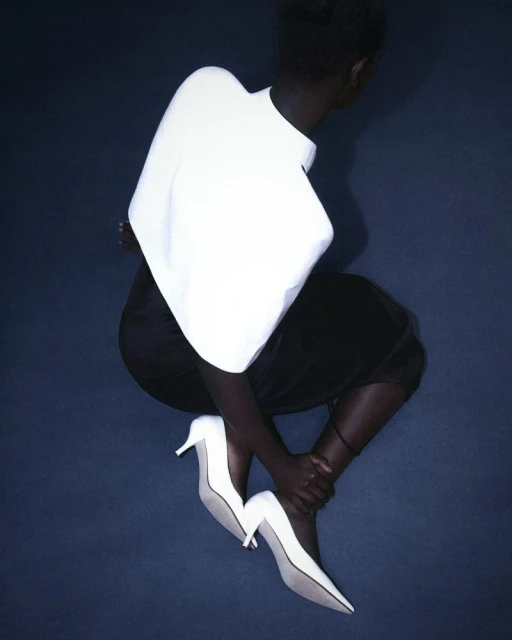 Fashion shot of a dark-skinned woman wearing a white shirt and black skirt, seated and holding one ankle while wearing pointed white heels.