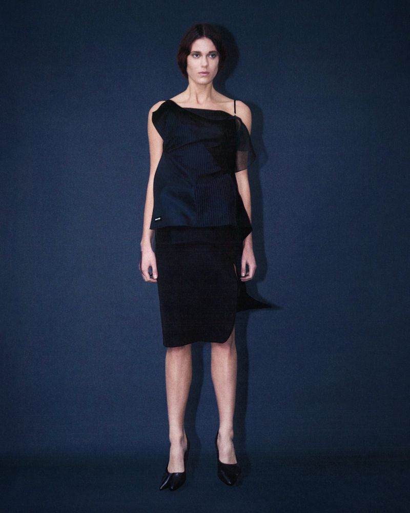 A female fashion model poses in a black asymmetrical top and pencil skirt against a blue background in a full shot in a studio.